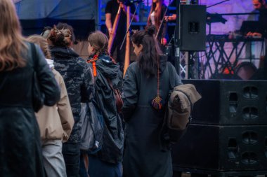 Yağmurluk ve ceket giyen çeşitli insanlar bir açık hava etkinliğinde toplanıyor; bazıları telefonlarını kontrol ediyor ve şemsiye taşıyor. Arkaplanda renkli kurdeleler asılı..