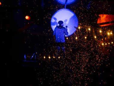 Bir sanatçı, koyu bir sahnede turuncu kıvılcımlarla çevrili, parlayan mavi bir davulun önünde duruyor ve güçlü bir görsel kontrast yaratıyor. Konser veya ayarları göster.