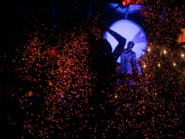 Bir sanatçı, karanlık bir sahnede turuncu kıvılcımlarla çevrili parlayan mavi bir davulun önünde duruyor ve güçlü bir görsel kontrast oluşturuyor. Bir konser ya da gösteri yeri.