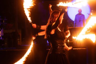 Kırmızı-turuncu alevler ve arka planda canlı bir grupla dramatik bir gece performansında iki ateş dansçısı alevli aksesuar çevirir..