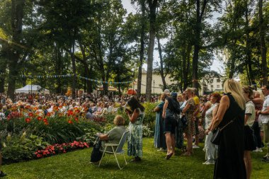 Ağaçlar ve çiçeklerle çevrili güneşli bir parkta, banklarda ve sandalyelerde oturan geniş bir izleyici bayrak altında açık hava etkinliğine katılıyor.. 