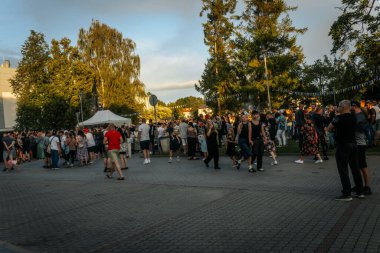 Sıcak ve güneşli bir günde, çocuklu aileler de dahil olmak üzere insanlar renkli asma kurdeleler ve çadırlar ile süslenmiş bir şenlik parkı yolunda yürürler..