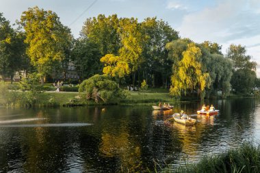 İnsanlar, sakin ve manzaralı bir yaz anının tadını çıkararak, altın akşam ışığında, ağaçların oluşturduğu göl yolu boyunca kürek çekerek gezerler. Valmiera, Letonya - 02.08.2025