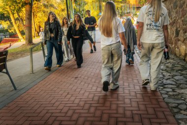Sıcak ve güneşli bir günde, çocuklu aileler de dahil olmak üzere insanlar renkli asma kurdeleler ve çadırlar ile süslenmiş bir şenlik parkı yolunda yürürler..