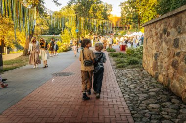 Sıcak ve güneşli bir günde, çocuklu aileler de dahil olmak üzere insanlar renkli asma kurdeleler ve çadırlar ile süslenmiş bir şenlik parkı yolunda yürürler..