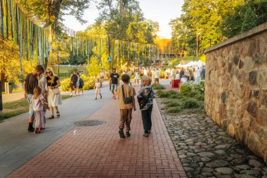 Sıcak ve güneşli bir günde, çocuklu aileler de dahil olmak üzere insanlar renkli asma kurdeleler ve çadırlar ile süslenmiş bir şenlik parkı yolunda yürürler..