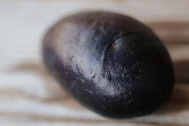 Ahşap bir yüzeye, yumuşak doğal ışık altında koyu tenli, çiğ, mor bir patatesin yakın çekim görüntüsü. Sığ alan derinliği ve görünür doku.