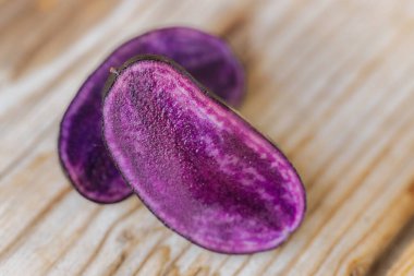 Mor bir patates diliminin makro çekimi yüzeyindeki parlak dokuyu ve nemi ahşap bir zemine karşı gösteriyor. Doğal ışıklandırma ve sığ alan derinliği.