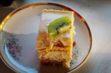 Bir dilim kremalı, katmanlı pasta ve bir dilim taze kivi. Dekoratif bir çiçek tabağında sıcak aydınlatmayla servis ediliyor..