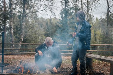 Bir adam ve bir çocuk dalları topluyor ve kırsal bir alanda ağaçlar ve banklarla çevrili bir ormanda yanan bir ateşe bakıyor..
