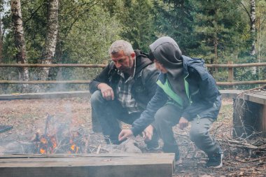 Bir adam ve bir çocuk dalları topluyor ve kırsal bir alanda ağaçlar ve banklarla çevrili bir ormanda yanan bir ateşe bakıyor..