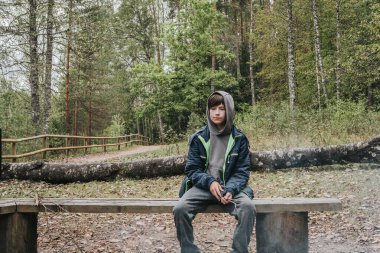 Gri kapüşonlu ve mavi ceketli bir çocuk sakince, ağaçtan bir bankta, sessiz bir ormanda, dumanlı bir kamp ateşinin altında, yeşil yapraklarla çevrili bir şekilde oturuyor..