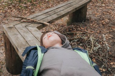 Gri kapüşonlu ve mavi ceketli genç bir çocuk ahşap bir yüzeye uzanmış, gözleri kapalı, rahat ya da ormanlık bir alanda uyuyor gibi görünüyor..
