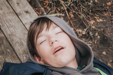 Gri kapüşonlu ve mavi ceketli genç bir çocuk ahşap bir yüzeye uzanmış, gözleri kapalı, rahat ya da ormanlık bir alanda uyuyor gibi görünüyor..