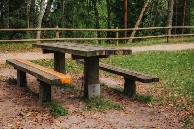 A worn-out wooden picnic table with benches, one of which has been partially renovated with fresh planks, is placed in a forest clearing near a dirt path and a wooden fence.