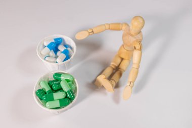A wooden mannequin sits on a white surface next to two paper cups filled with green and blue capsules. A conceptual health or choice theme.