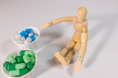 A wooden mannequin sits on a white surface next to two paper cups filled with green and blue capsules. A conceptual health or choice theme.