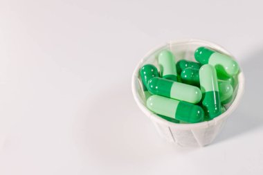 Paper cup filled with green capsules placed on a white surface with enough space for text. Medical or healthcare related concept.