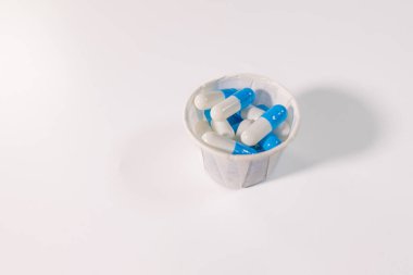 Beyaz bir yüzeye yerleştirilmiş mavi ve beyaz kapsüllerle dolu küçük bir kağıt bardak ve metin için bolca yer var. Tıbbi ya da ilaç teması.