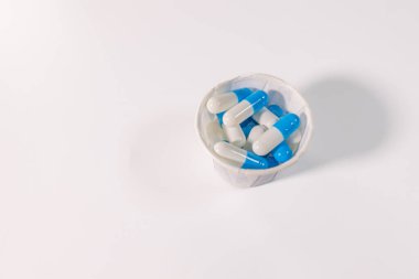 Beyaz bir yüzeye yerleştirilmiş mavi ve beyaz kapsüllerle dolu küçük bir kağıt bardak ve metin için bolca yer var. Tıbbi ya da ilaç teması.