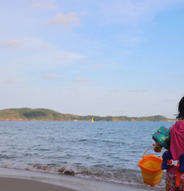 Küçük bir kız elinde plastik bir kum kovası tutuyor, Tayland, Rayong sahilinde duruyor.