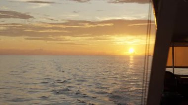 Bir tekneden çekilen muhteşem gün batımı görüntüsü, sıcak turuncu ve pembe renkler ile gökyüzünü boyuyor. Sakin sular göksel tuvali yansıtıyor ve nefes kesici bir manzara yaratıyor. Yüksek kalite 4k görüntü