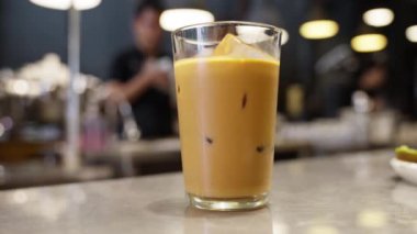 Bir kafede nefis bir buzlu kahvenin sunulduğu bir davet sahnesi. Meyve suyu ustası barista arka planda yürüyor, enerji ve işçilik elementi kahve dükkanının atmosferini geriletiyor. 