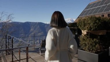 Lüks bir tatil sahnesi: Bornozlu kadın görkemli kar örtülü dağlara bakıyor. Huzurlu bir ortam ve rahat bir kıyafetle, doğanın güzelliğine kendini kaptırıyor, rahatlamanın somut bir örneği..