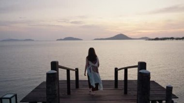 Bir kadının, batan güneşin parlak renkleriyle çerçevelenmiş bir köprüde zarif bir şekilde yürüdüğü simetrik bir görüntü. Aşağıdaki suda yansıyan yansımalarla, güzelliğin tesellisiyle. Yüksek kalite 4k görüntü