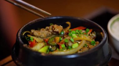 Siyah taştan bir kasede Kore usulü kızarmış pilavın yakın çekimi. Çubuklar taze sığır eti ve renkli sebzeleri topluyor. Cızırtılı sunumu ve lezzetli aromayı vurguluyorum. Yüksek kalite 4k görüntü