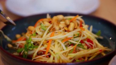 Tayland papaya salatasına yakın çekim, canlı renkler ve doğranmış yeşil papaya, vişneli domates ve fıstık gibi taze malzemeler. Keskin, baharatlı aromalar ve taze dokular vurgulanıyor..