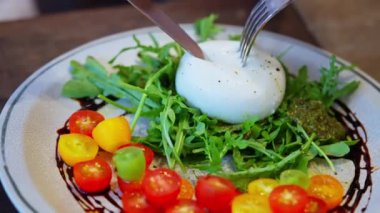 Bıçak ve çatalla kesilen burrata salatasının yakın plan çekimi, içinde olgun vişneli domateslerin üzerine sızan kremalı burrata var. Taze, zengin dokuları ve canlı renkleri vurgulamak. Yüksek kalite 4k görüntü
