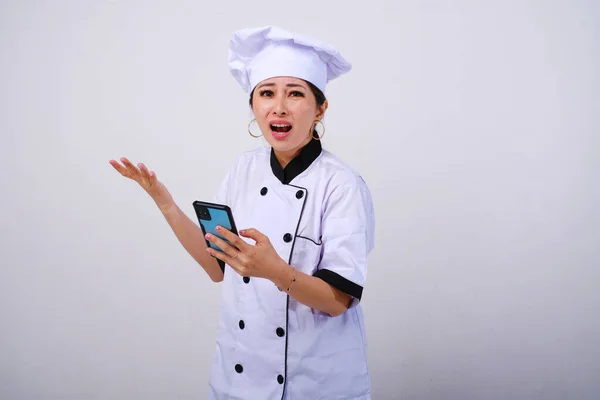 Photo of disappointed and confused female Asian chef using mobile phone reacting to bad review on isolated white background