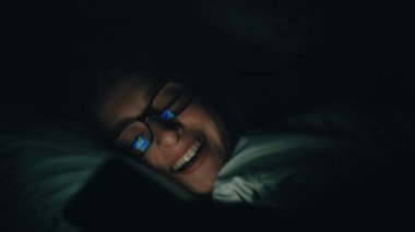 Yatakta cep telefonu kullanan güzel beyaz kadın, gülüyor, karanlık arka plan. ARRI Alexa ile 120 fps hızla ArriRaw 'da çekilmiş, 23.98 fps QuickTime Apple ProRes 422 HQ ihraç edilmiş..