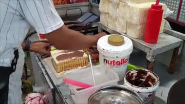 Klungkung, Bali, Endonezya - 29 Mart 2024: Klungkung Bali pazarında Nutella markalı tatlı fındık ezmesi ile beyaz ekmek yayan bir tost satıcısı.
