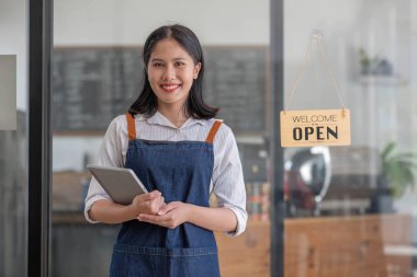 Önlüklü güzel ve arkadaş canlısı Asyalı bir bayan garson elinde dijital tabletle kafenin girişinde duruyor. Konukseverlik hizmeti, iş girişimcisi, küçük işletme girişimi..