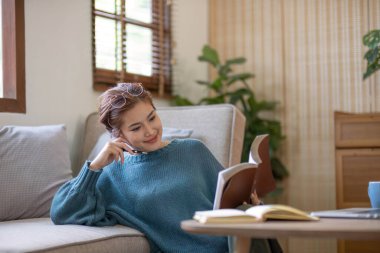 Evdeki rahat gri koltukta kitap okuyan genç bir kadın...