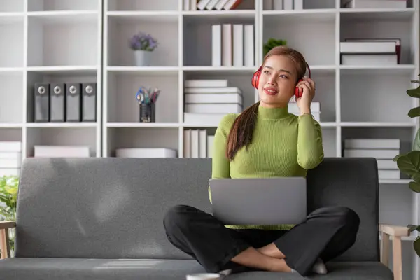 Happy and relaxed young Asian woman listening to music through her headphones on a sofa in her living room