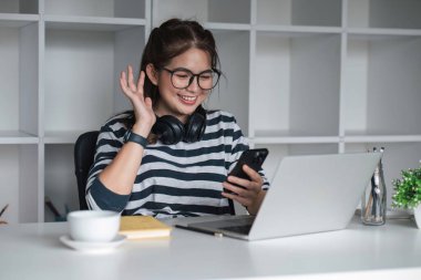 Asyalı üniversite öğrencisi çalışırken bilgisayar ve kulaklıkla telefon kullanıyor. Video çağrısı ile mesajlar okunuyor ve arkadaşlara selam veriliyor.