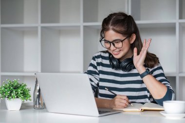 Asyalı kız öğrenci çevrimiçi sınıfta çalışıyor, çevrimiçi çalışıyor, kulaklık takıyor, video izliyor, zumluyor, bilgisayarlı dizüstü bilgisayarla mutlu Asyalı kadın öğrenme dili öğreniyor...