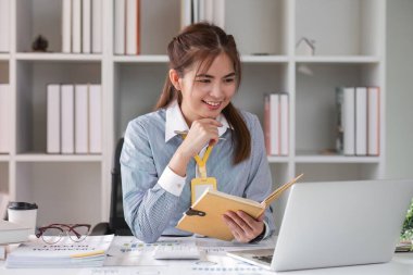 Ofiste dizüstü bilgisayar kullanırken not alan bir iş kadını. Kadın girişimci, dizüstü bilgisayarla çalışırken ayrıntıları kağıda yazıyor. Gülümseyen Asyalı iş kadını.