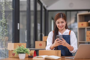 Arka planda paketlerle dolu ofiste oturan genç bir Asyalı kadının portresi. Emirleri yaz ve KOBİ iş ekomünyesi ve teslimatı için akıllı telefon kullan..