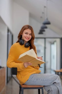 Okul kütüphanesinde elinde fotokopi kitabı tutarak gülümseyen kız öğrenci evden uzaktan ev ödevini internetten hazırla..