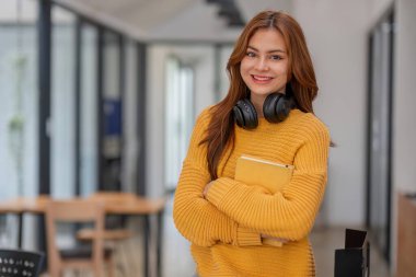 Okul kütüphanesinde elinde fotokopi kitabı tutarak gülümseyen kız öğrenci evden uzaktan ev ödevini internetten hazırla..