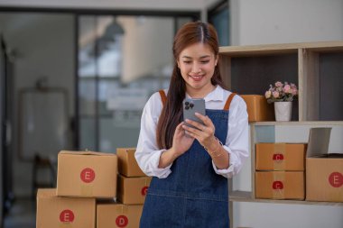 Arka planda paketlerle dolu bir ofiste duran genç bir Asyalı kadının portresi. Emirleri yaz ve KOBİ için akıllı telefon kullan..