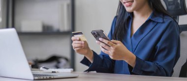 Akıllı telefon ve kredi kartını elinde tutan çekici Asyalı kadın, mobil bankacılık uygulaması veya çevrimiçi alışveriş uygulaması kullanıyor...