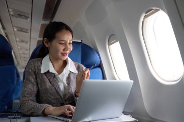 Uçaktaki çekici Asyalı kadın yolcu dizüstü bilgisayar ve kredi kartı kullanarak rahat koltukta oturuyor, kablosuz bağlantıyı kullanarak alışveriş yapıyor, tarz olarak seyahat ediyor..