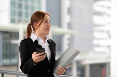 Takım elbiseli genç Asyalı iş kadınının portresi modern ofis binası konseptinin ve insanların önünde dururken akıllı telefondan konuşuyor..