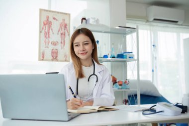 Steteskoplu beyaz tıbbi üniformalı genç Asyalı doktor sağlık kliniğinde ya da hastanede hastalarla konuşurken dizüstü bilgisayar görüntüsü kullanıyor..