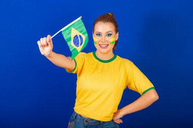 Beyaz kadın, kızıl saçlı, Brezilyalı futbol fanatiği, Brezilyalı, mavi arka plan, Brezilya bayrağı sallayan.